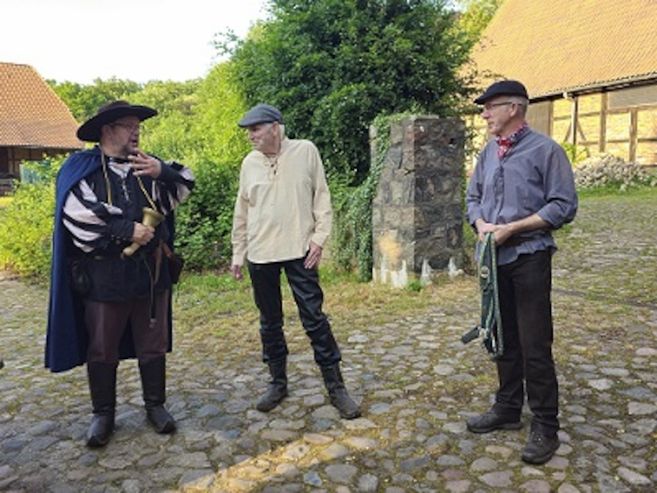 Drei Männer in historischer Kleidung stehen auf einem gepflasterten Hof vor alten Gebäuden.