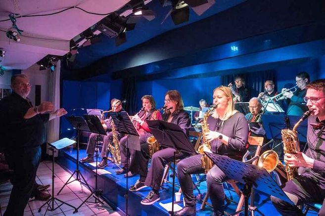 Eine Big Band spielt auf einer Bühne, dirigiert von einem Mann. Die Musiker spielen Saxophone und Trompeten.