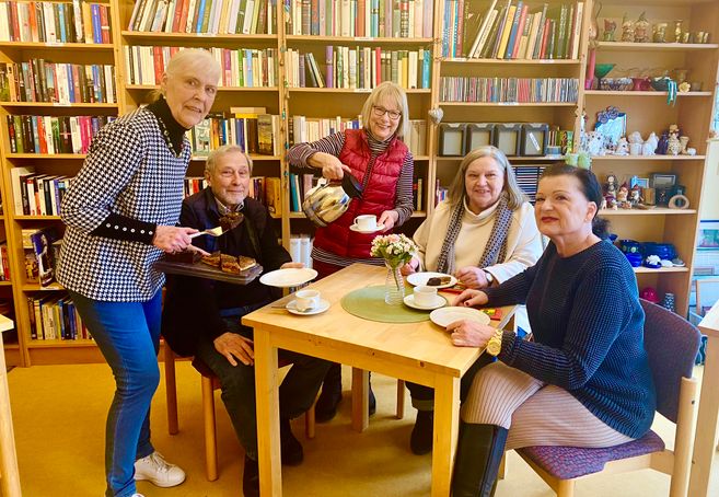 Eine Gruppe älterer Menschen sitzt in einem Café mit Bücherregalen im Hintergrund. Eine Frau serviert Kuchen und Kaffee.