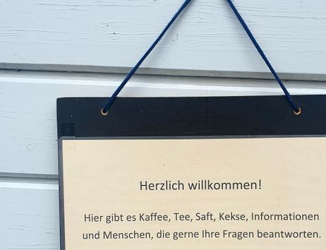 Ein Schild an einer Wand mit einer Einladung zu Kaffee, Tee, Saft und Keksen sowie Informationen und freundlichen Menschen.