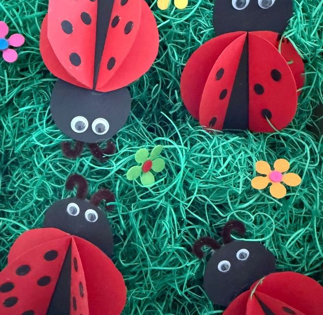 Bastelarbeiten von Marienkäfern aus Papier auf grünem Hintergrund mit bunten Blumen.