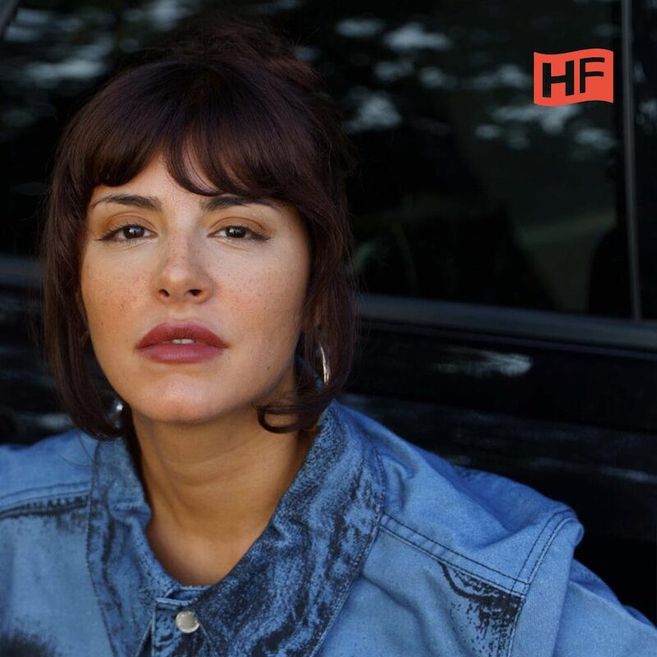 Frau mit braunen Haaren und Jeansjacke vor einem dunklen Hintergrund mit HF-Logo.