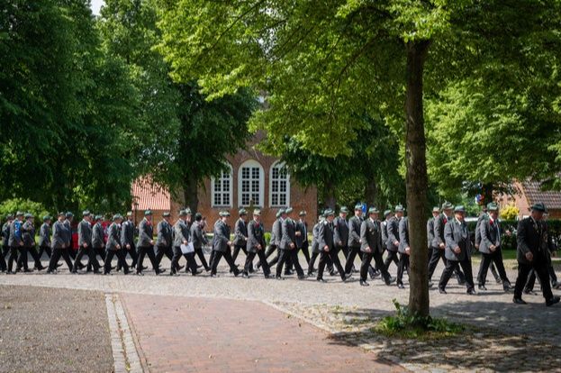 Eine Gruppe von Menschen in Uniformen marschiert auf einem gepflasterten Weg, umgeben von Bäumen und einem Backsteingebäude im Hintergrund.