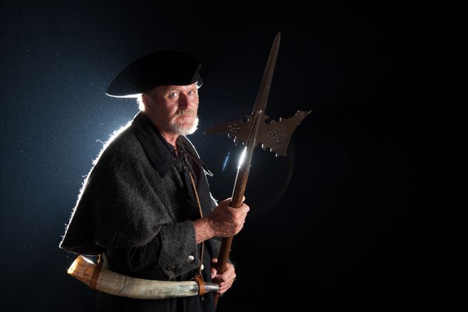 Ein Mann in historischer Nachtwächterkleidung mit Hellebarde und Horn, vor dunklem Hintergrund.