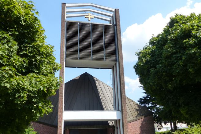 Moderne Kirche mit markantem Turm und Kreuz, umgeben von Bäumen.