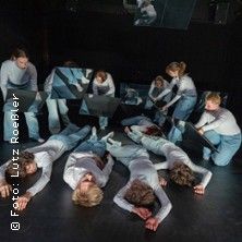 Eine Gruppe von Menschen in weißen und blauen Kostümen liegt und steht auf einer Bühne, umgeben von Spiegeln.