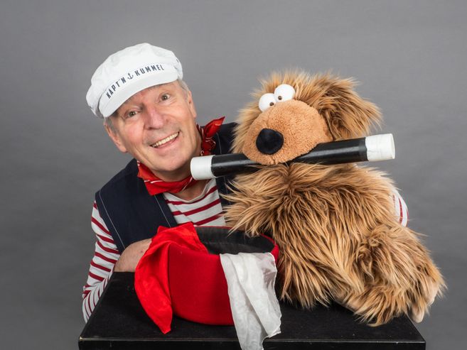 Ein Mann in Matrosenkleidung mit einer weißen Mütze und ein Plüschtier mit einem Zauberstab im Mund.