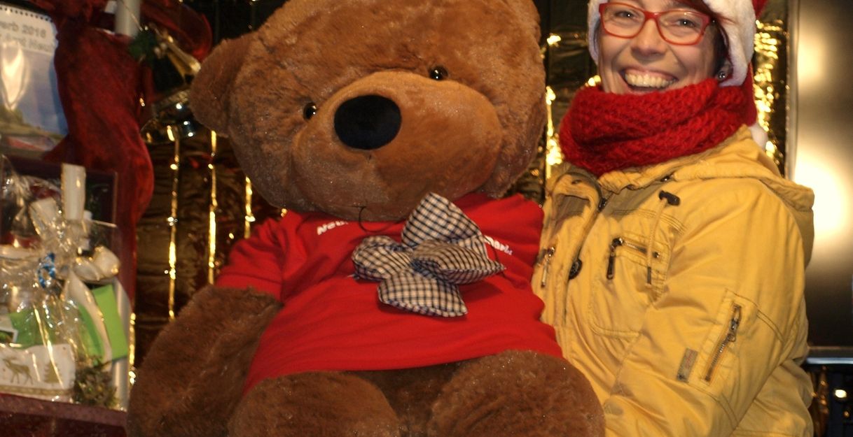 Eine Person mit Weihnachtsmütze hält einen großen Plüschbären auf einem Weihnachtsmarkt.