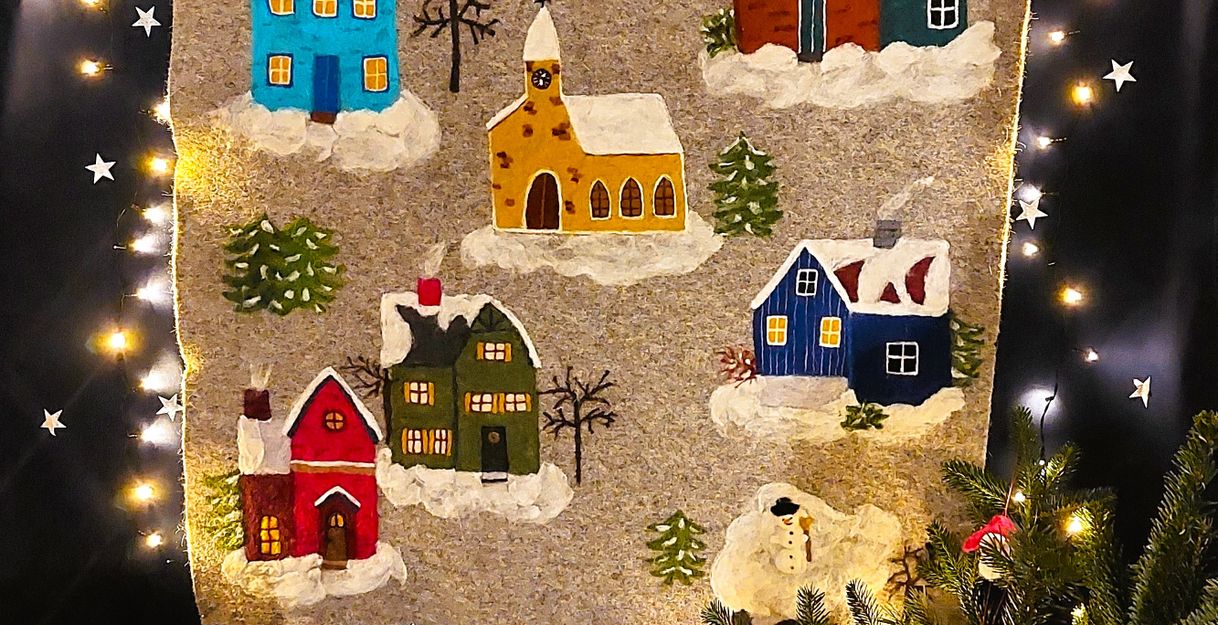 gefilzter Wandbehang mit schneebedeckten, bunten Häusern, umgeben von einer Lichterkette