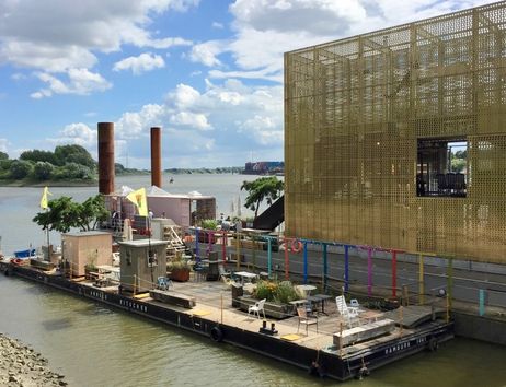 Schwimmende Plattform mit Sitzgelegenheiten und Pflanzen am Fluss, daneben modernes Gebäude mit goldener Fassade.