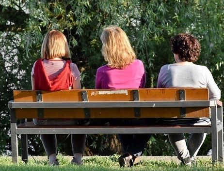 Drei Personen sitzen auf einer Parkbank, den Betrachtenden den Rücken zugewandt. 