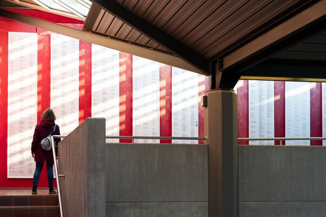 Eine Person betrachtet eine Wand mit langen Listen von Namen, die auf weißen Tafeln vor einem roten Hintergrund angebracht sind.
