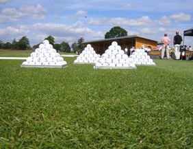Golfbälle in Pyramidenform auf einer Driving Range mit Personen im Hintergrund.