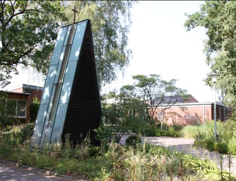 Ein modernes, dreieckiges Kirchturmgebäude mit einem Kreuz an der Spitze, umgeben von Bäumen und Pflanzen, mit einem Backsteingebäude im Hintergrund.