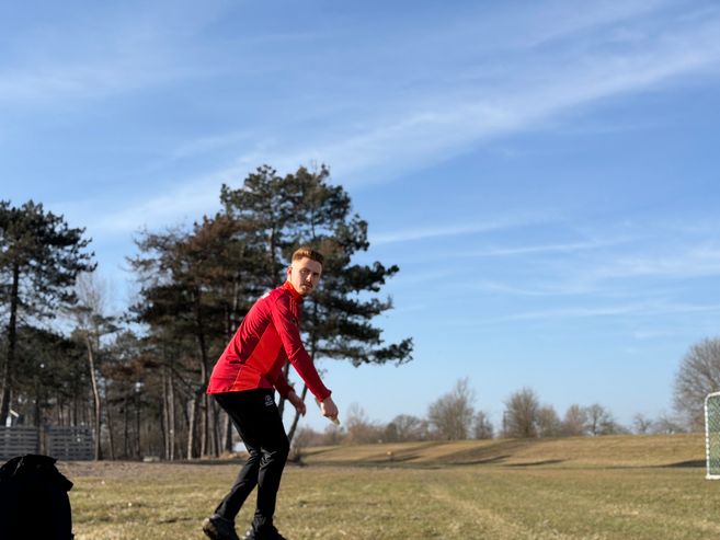 Ein Mann in roter Sportkleidung spielt Discgolf auf einem offenen Feld mit Bäumen im Hintergrund.