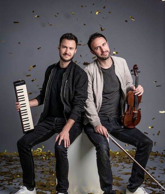 Zwei Männer sitzen auf einem Hocker, umgeben von goldenen Konfetti. Einer hält eine Melodica, der andere eine Geige.