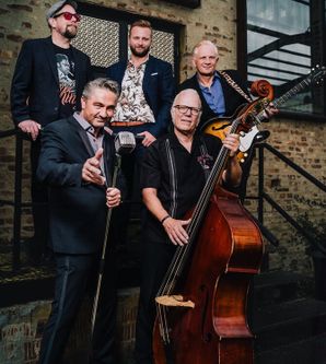 Eine fünfköpfige Band posiert mit Instrumenten vor einem Backsteingebäude.