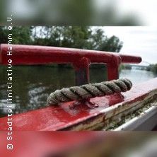 Nahaufnahme eines roten Geländers auf einem Boot mit einem Seil, das um das Geländer gewickelt ist. Im Hintergrund ist ein Fluss und Bäume zu sehen.