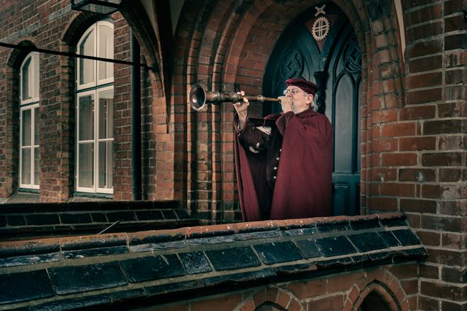 Ein Mann in historischer Kleidung bläst ein Horn auf einem Balkon vor einem Backsteingebäude.