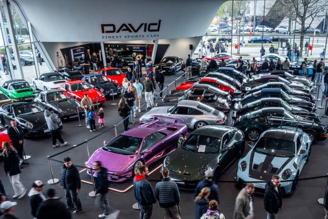 Zahlreiche Sportwagen und Oldtimer, darunter Porsche-Modelle und ein lila Supersportwagen, stehen dicht an dicht vor dem Showroom von DAVID Finest Sports Cars, umgeben von Besucherinnen und Besuchern.
