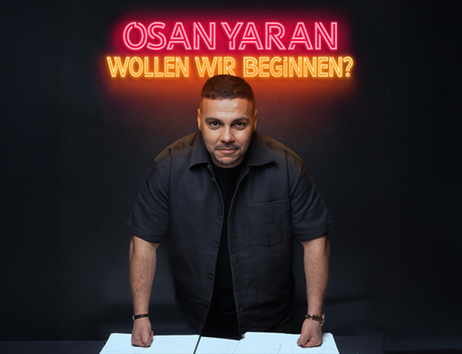 Ein Mann steht vor einem Tisch mit Neon-Schriftzug 'Osan Yaran - Wollen wir beginnen?' im Hintergrund.