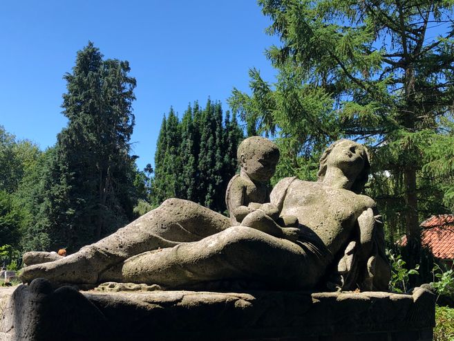 Steinskulptur einer liegenden Frau mit Kind im Freien, umgeben von Bäumen.