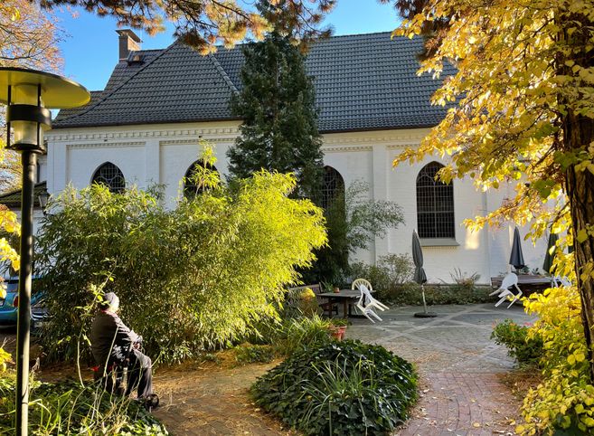 Ein ruhiger Garten mit einer weißen Kirche im Hintergrund, umgeben von Bäumen und Pflanzen. Eine Person sitzt auf einer Bank im Vordergrund.