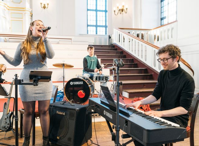 Eine Jugendband probt in einer Kirche. Eine Sängerin singt, während ein Schlagzeuger und ein Keyboardspieler musizieren.