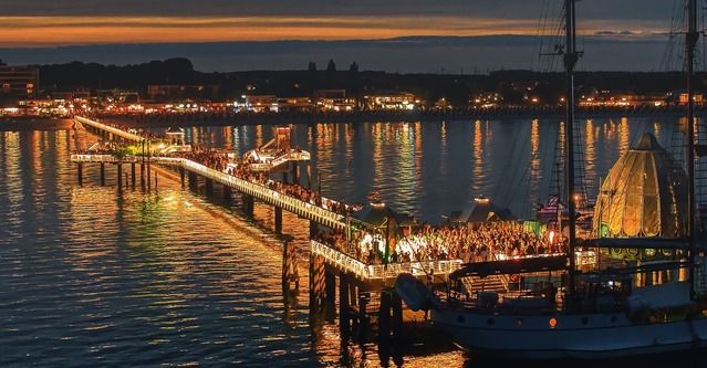 Eine beleuchtete Seebrücke bei Nacht mit Menschenmengen und einem Schiff im Vordergrund.