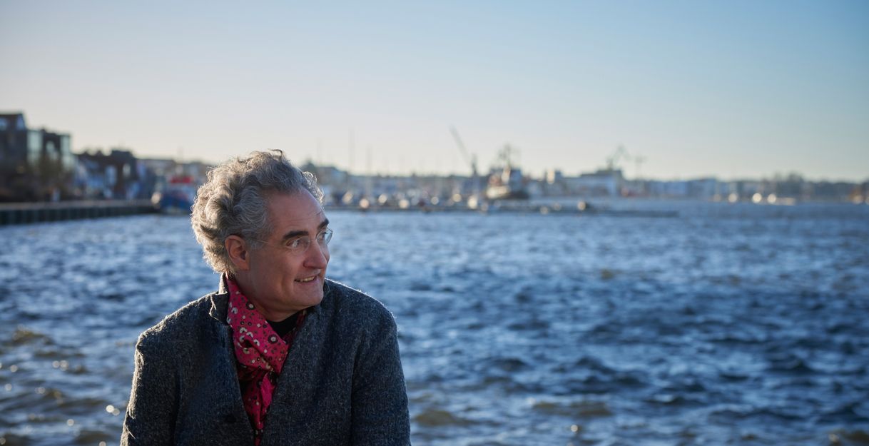 Ein Mann mit grauen Haaren und Brille sitzt am Wasser, im Hintergrund ist eine Stadtlandschaft mit Kränen zu sehen.