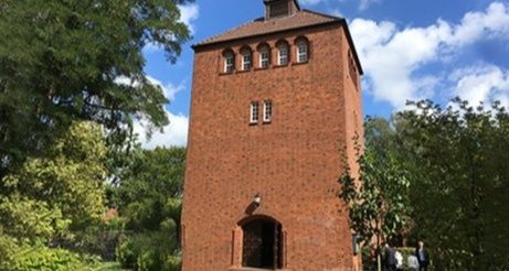 Backsteinturm mit Bogenfenstern und Eingang, umgeben von Bäumen, unter blauem Himmel.