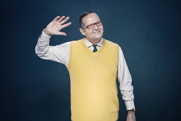 Ein Mann in gelbem Pullunder winkt und lächelt vor blauem Hintergrund.