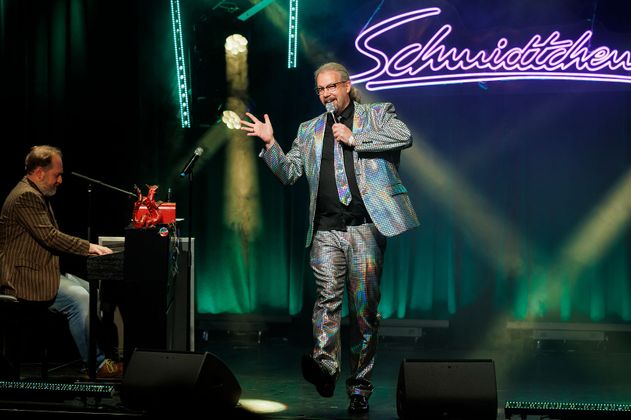 Ein Mann in einem glitzernden Anzug singt auf einer Bühne, während ein anderer Mann Klavier spielt. Im Hintergrund leuchtet ein Schild mit der Aufschrift 'Schmidtchen'.