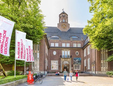 Menschen vor einem palastähnlichen Gebäude. Der rote Backsteinbau geht über 5 Etagen. Der Eingangsbereich ist über Treppen erreichbar, hat ein steinernes Tor und ist von Fenstern umrandet. Darüber ein Balkon. Auf dem Dach ist mittig ein Turm.