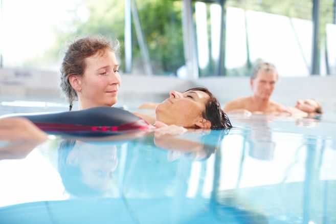 Zwei Personen im Wasser, eine hält die andere in einer entspannten Position. Im Hintergrund ein weiteres Paar in ähnlicher Pose.