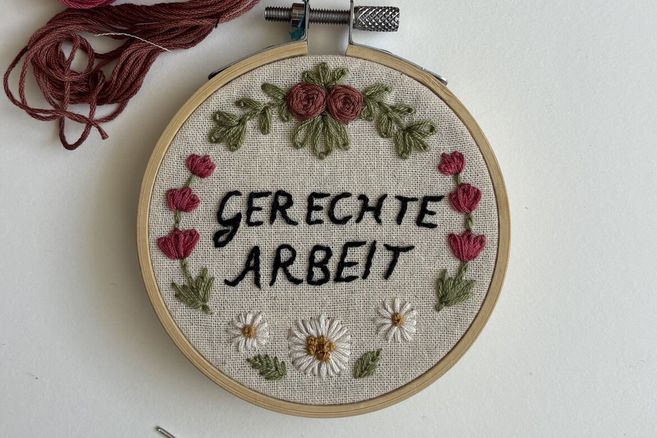 Ein Stickrahmen mit der Aufschrift 'Gerechte Arbeit', umgeben von gestickten Blumen und Blättern.