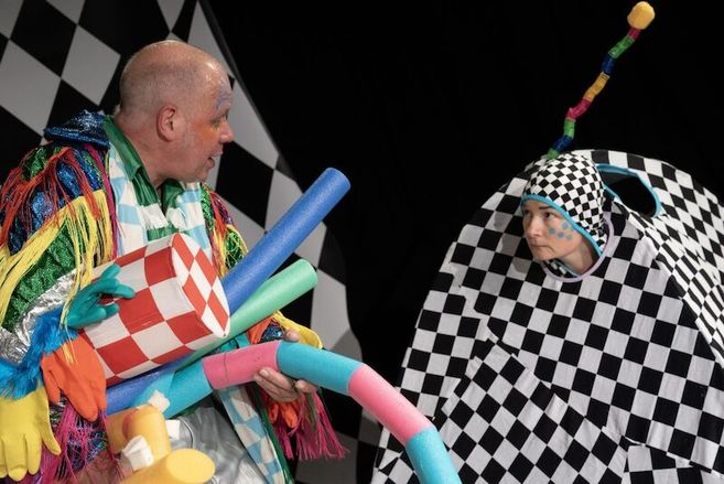 Zwei Schauspieler in bunten Kostümen auf einer Bühne mit Schachbrettmuster-Hintergrund.