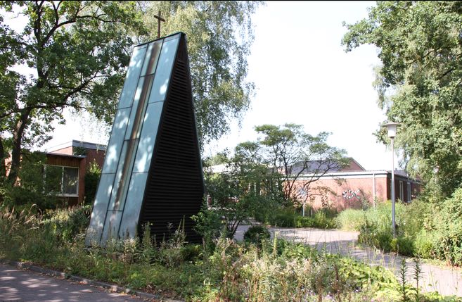 Ein modernes, dreieckiges Kirchturmgebäude mit einem Kreuz an der Spitze, umgeben von Bäumen und Pflanzen, mit einem Backsteingebäude im Hintergrund.