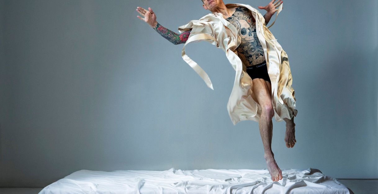 A performer in a cream silk robe and black underwear leaps from a white bed, his torso intricately tattooed.