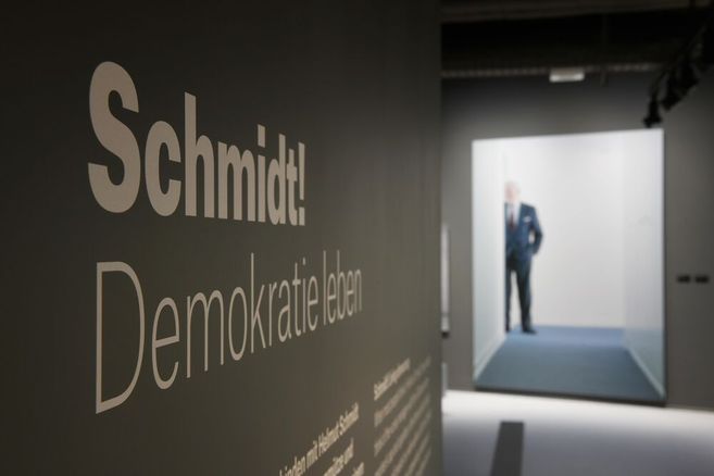 Fotos, das eine von der Seite aufgenommene Informationstafel im Vordergrund zeigt. Darauf ist deutlich "Schmidt! Demokratie leben" zu lesen. Im Hintergrund steht hinter einem Durchgang ein Mann im Anzug, der nur verschwommen zu sehen ist.