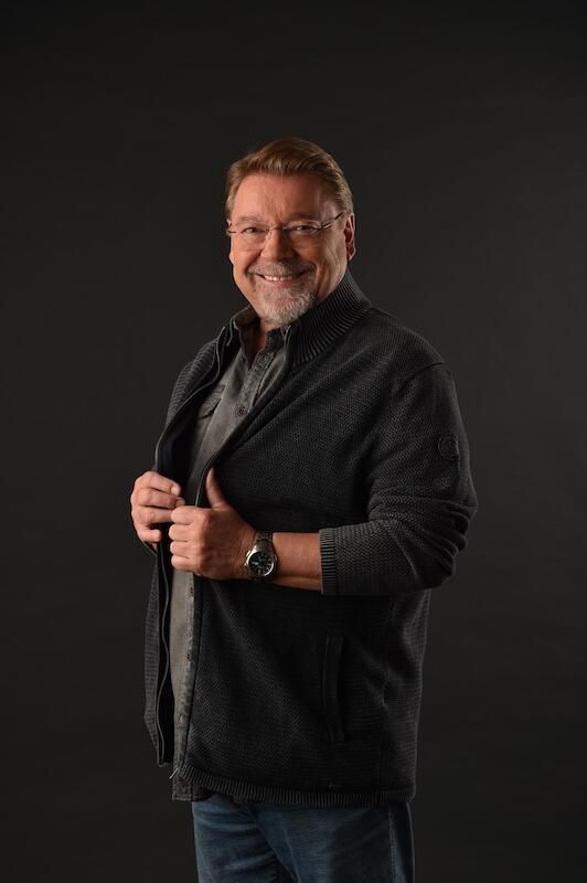 Ein lächelnder Mann in dunkler Kleidung vor einem schwarzen Hintergrund.