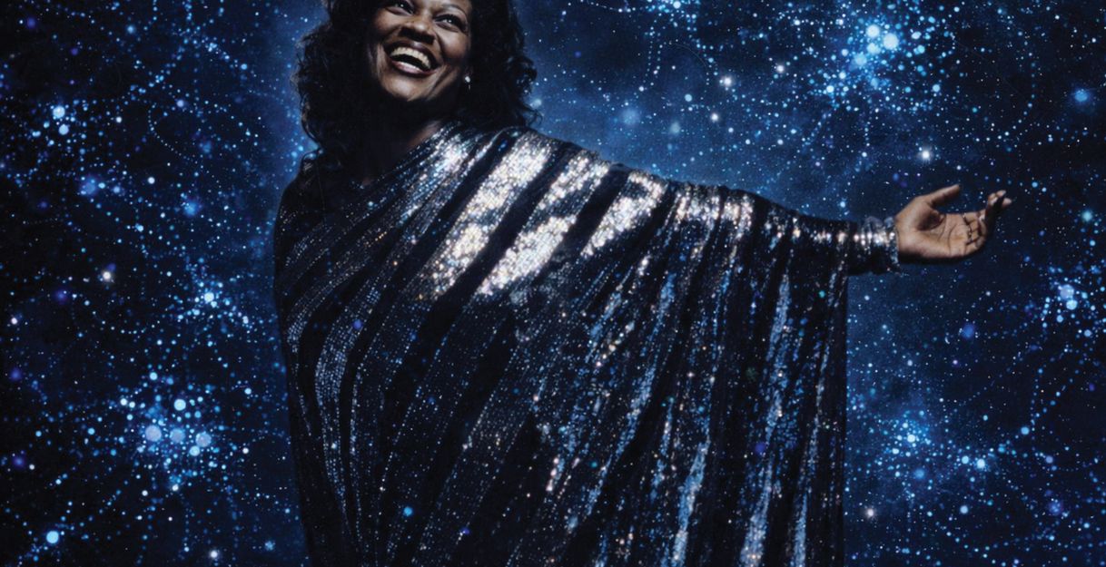 A woman stands in a glittering silver dress against the backdrop of a dark blue surface covered with many lights.