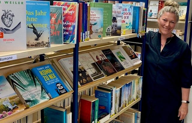 Eine Frau steht lächelnd neben Bücherregalen in einer Bücherei.