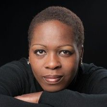 Porträt einer Person mit kurzem Haar und schwarzem Oberteil vor schwarzem Hintergrund.