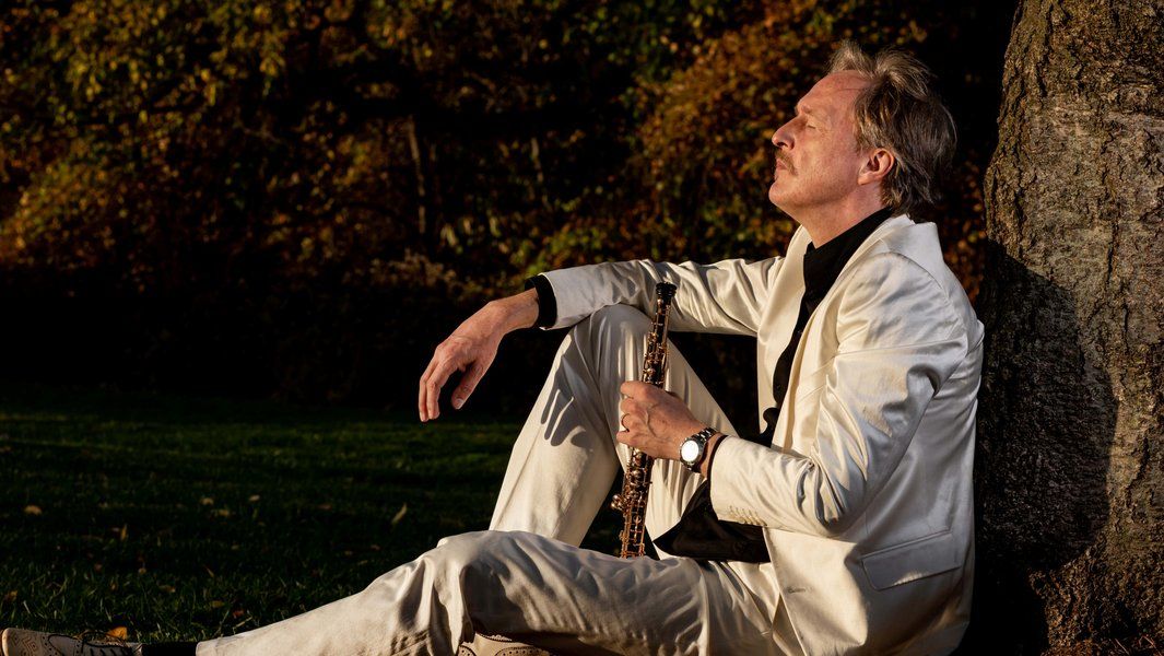 Ein Mann in einem weißen Anzug sitzt entspannt an einem Baum und hält ein Musikinstrument. Die Umgebung ist herbstlich mit buntem Laub.