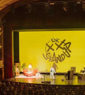 Ein Stand-up-Comedian auf einer Bühne in einem prunkvollen Theater mit gelbem Hintergrund und der Aufschrift 'KDK Stand Up'.
