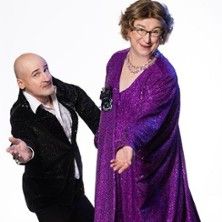 Zwei Personen in glamouröser Kleidung, eine in einem lila Kleid, die andere in einem schwarzen Anzug, posieren fröhlich vor einem weißen Hintergrund.