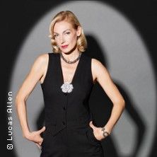 Eine Frau in eleganter schwarzer Kleidung steht vor einem grauen Hintergrund.