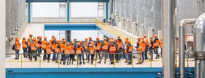 Gruppe von Menschen in orangefarbenen Sicherheitswesten und Helmen in einer Fabrikhalle.