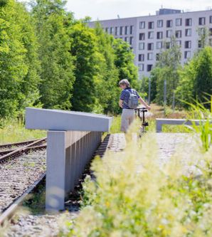 Blick aus Kniehöhe. Links sind zwei Gleise zu sehen. Mittig eine lange Reihe von Informationstafeln aus Beton, die von einem Menschen mit Fahrrade betrachtet werden. Im Hintergrund sind grüne Bäume sowie eine Reihenhauszeile zu sehen.